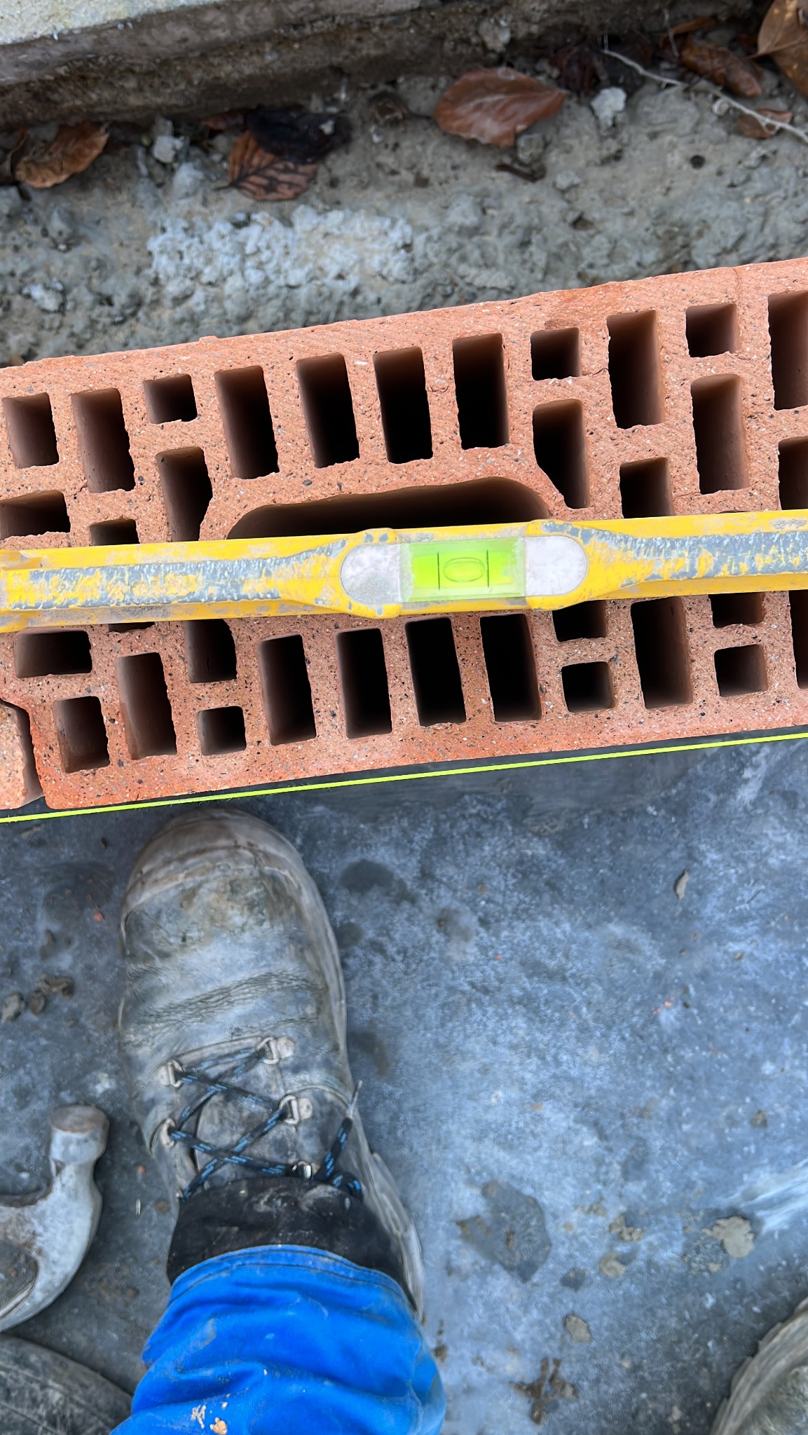 Cegła dziurawka poziomowana żółtą poziomicą, widoczny fragment buta roboczego murarza i młotek