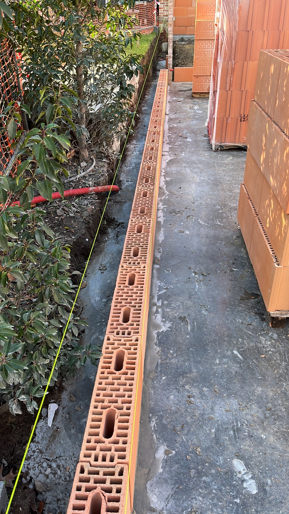 Wąski wykop z ułożonym pierwszym rzędem bloczków ceramicznych z pustakami, wyznaczony żółtą linką, przygotowany pod fundament budynku.