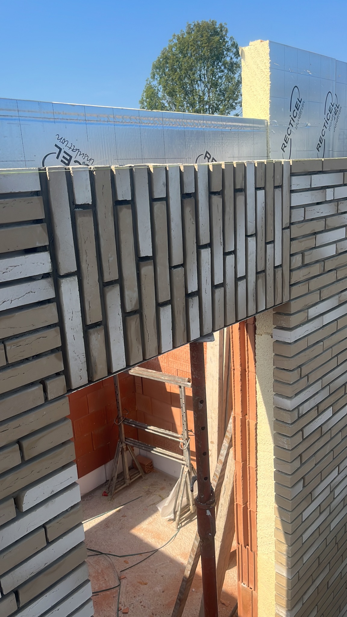 Wznoszenie ściany z cegły klinkierowej w jasnych odcieniach, widoczna izolacja termiczna i rusztowanie wewnątrz budynku.