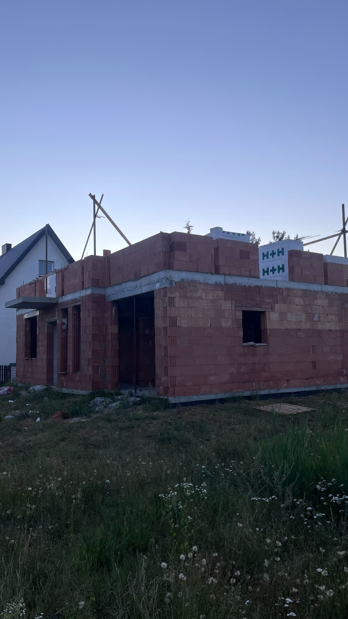 Surowy stan ścian domu z czerwonej cegły H+H, z widoczną konstrukcją stropu i otworami okiennymi, na tle nieba o zmierzchu i sąsiedniego domu z białymi ścianami i ciemnym dachem.