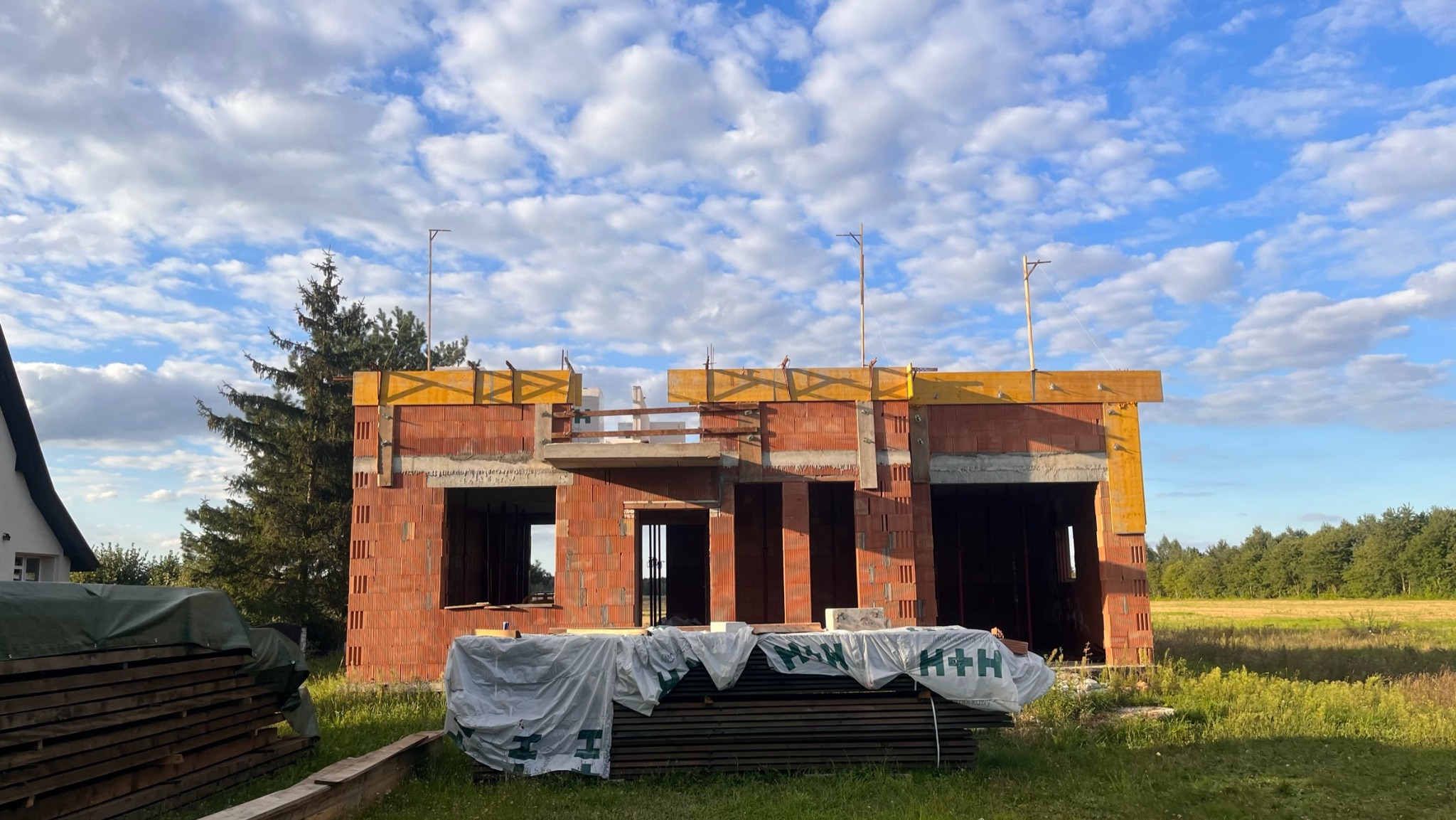 Wznoszenie ścian domu jednorodzinnego z czerwonej cegły, widoczne żółte elementy konstrukcyjne stropu, w tle błękitne niebo z chmurami, na pierwszym planie deski przykryte folią.