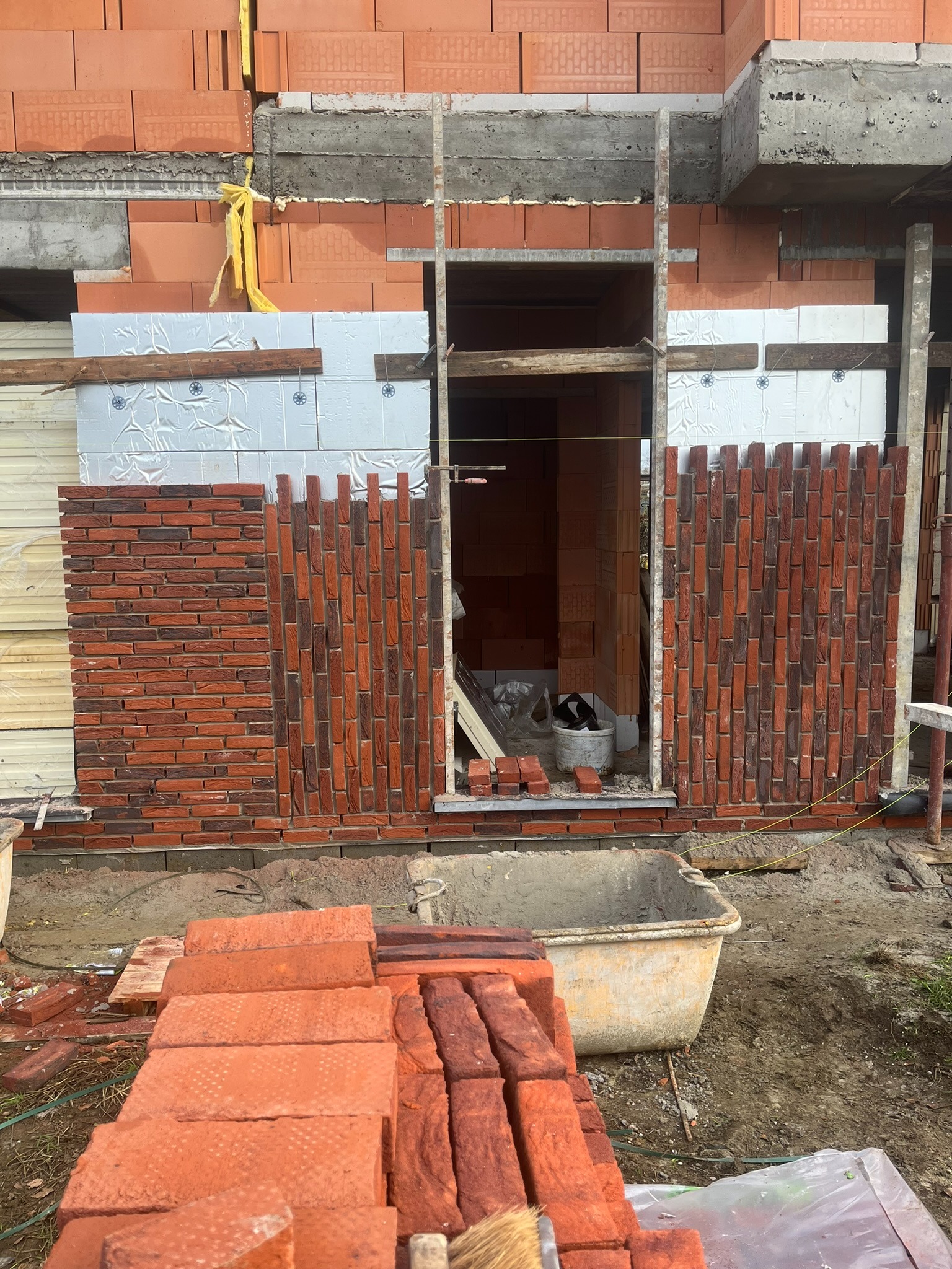Częściowo wymurowana ściana z czerwonej cegły klinkierowej, widoczne ocieplenie budynku i przygotowane cegły na ziemi, w tle wnętrze budynku w trakcie budowy.