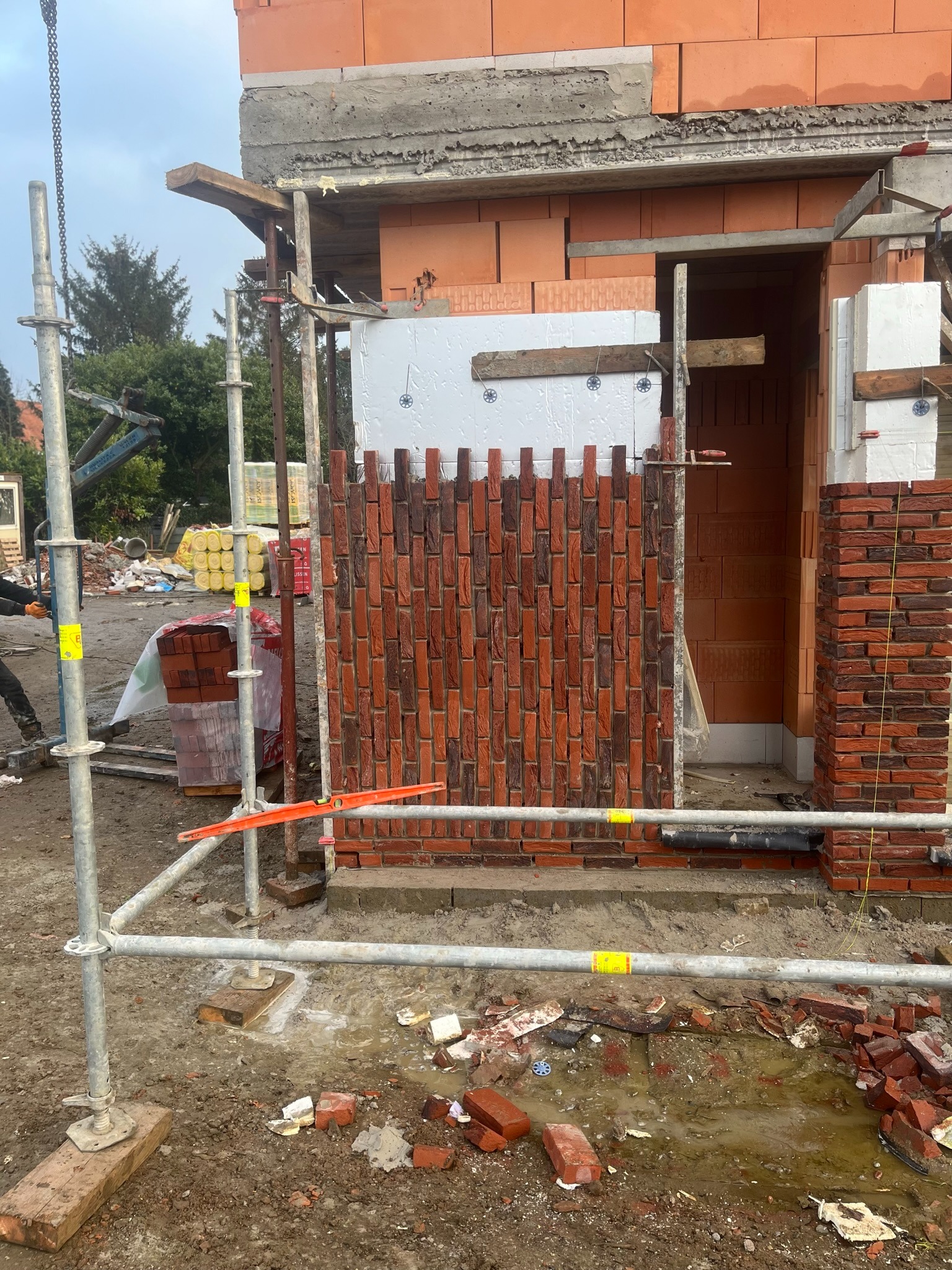 Wznoszenie ściany z czerwonej cegły klinkierowej na nowym budynku, widoczne rusztowanie i izolacja.