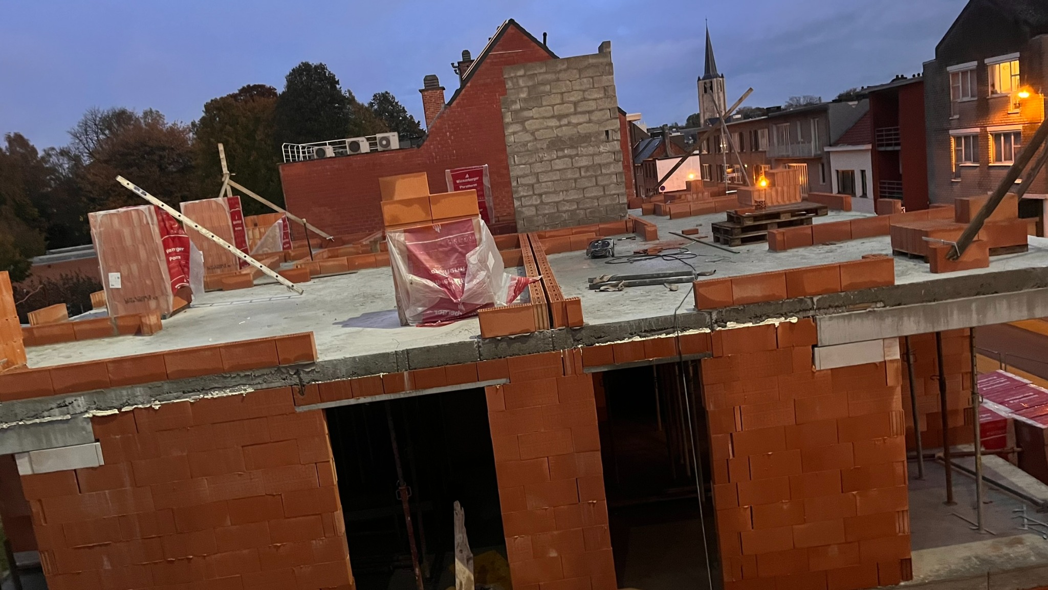 Wznoszenie ścian z czerwonej cegły na budowie, widok z góry na pustaki, zaprawy i narzędzia, w tle zabudowa miejska i kościół.