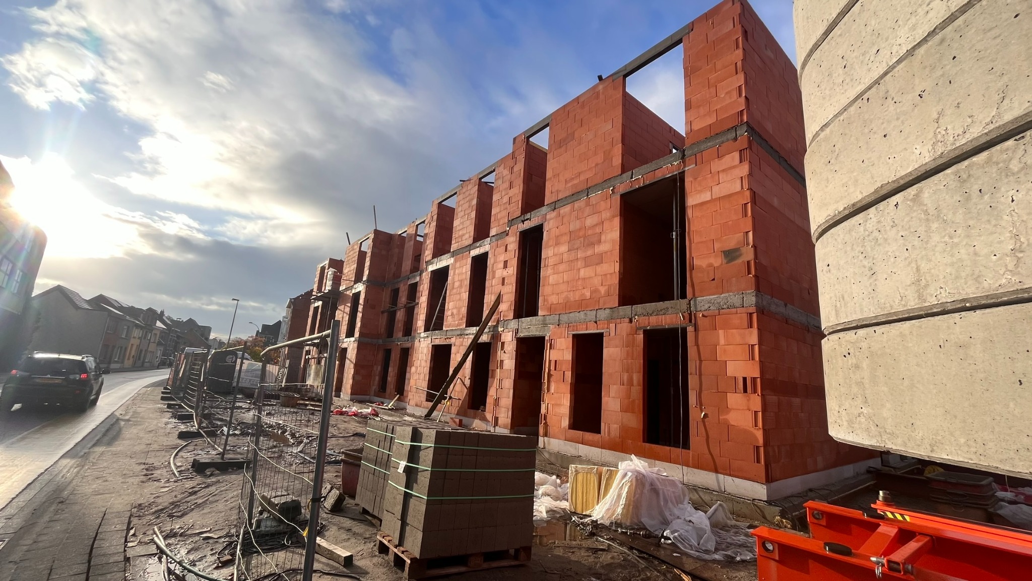 Budowa szeregowca z czerwonej cegły: widok na wznoszone ściany z otworami okiennymi, palety z cegłami na placu budowy, fragment betonowego elementu konstrukcyjnego w kadrze.
