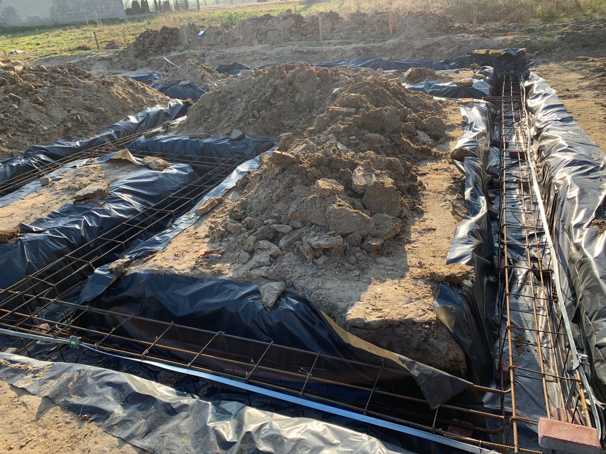 Przygotowanie wykopu pod fundamenty z izolacją folią i zbrojeniem stalowym, widoczny nasyp ziemi i kamieni.