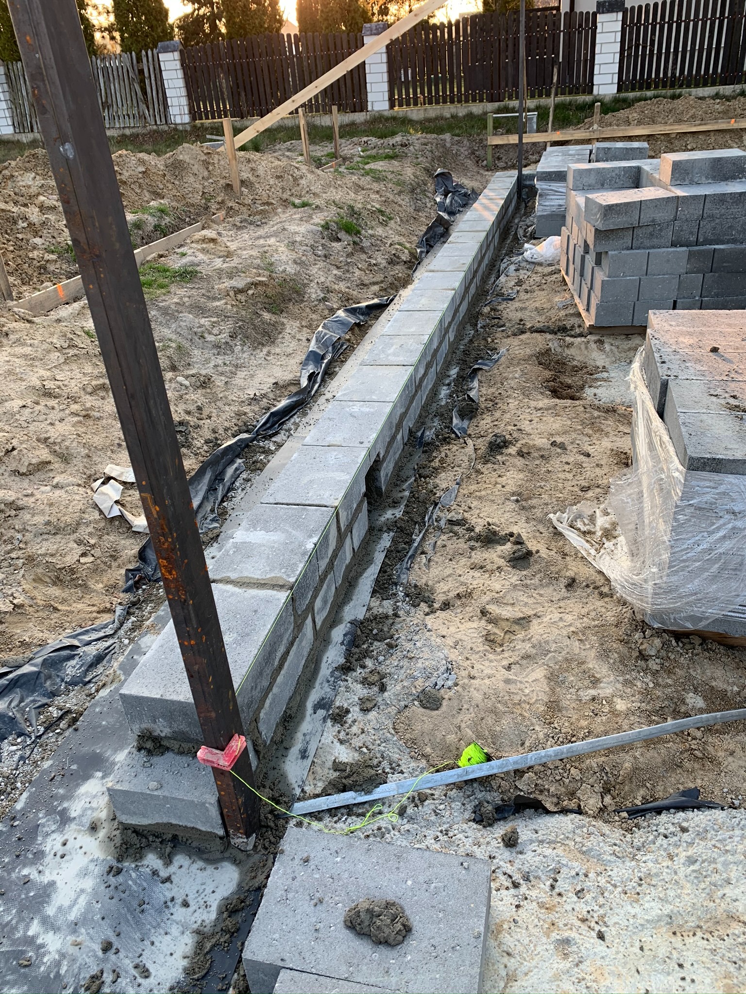 Wznoszenie fundamentu z betonowych bloczków z użyciem stalowego słupka i sznurka murarskiego, widoczna izolacja przeciwwilgociowa i zapas bloczków na budowie.
