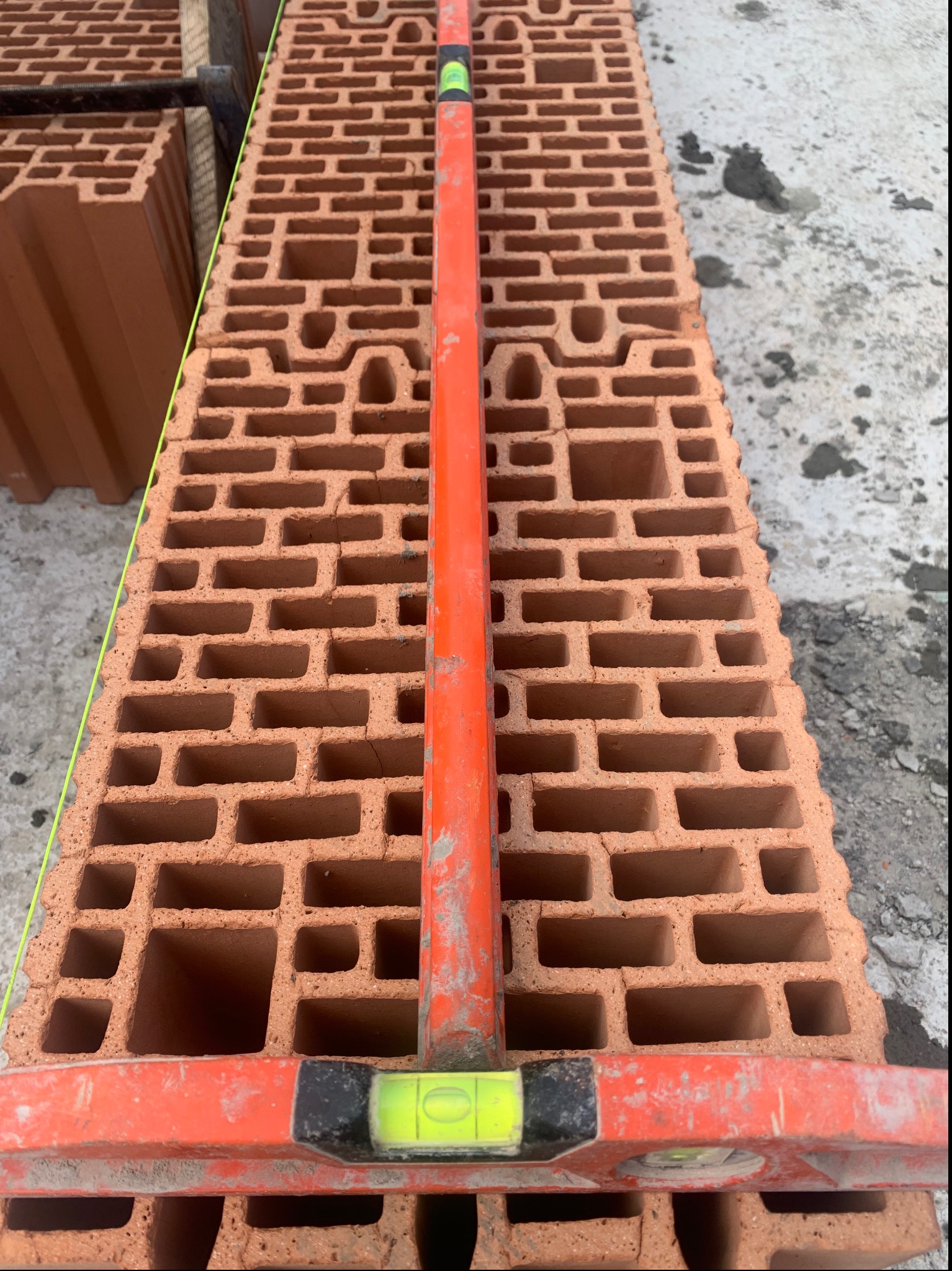 Ułożony warstwami bloczek ceramiczny z pustakami, sprawdzany poziomicą w kolorze pomarańczowym, z widoczną zieloną linką murarską, na tle betonowej powierzchni.