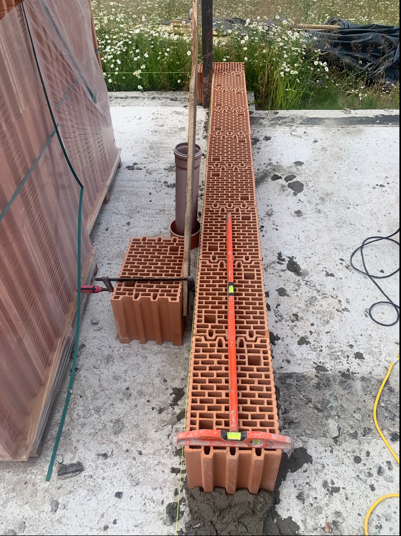 Wznoszenie ściany z pustaków ceramicznych Porotherm z użyciem poziomicy murarskiej, na tle łąki z kwitnącymi stokrotkami.