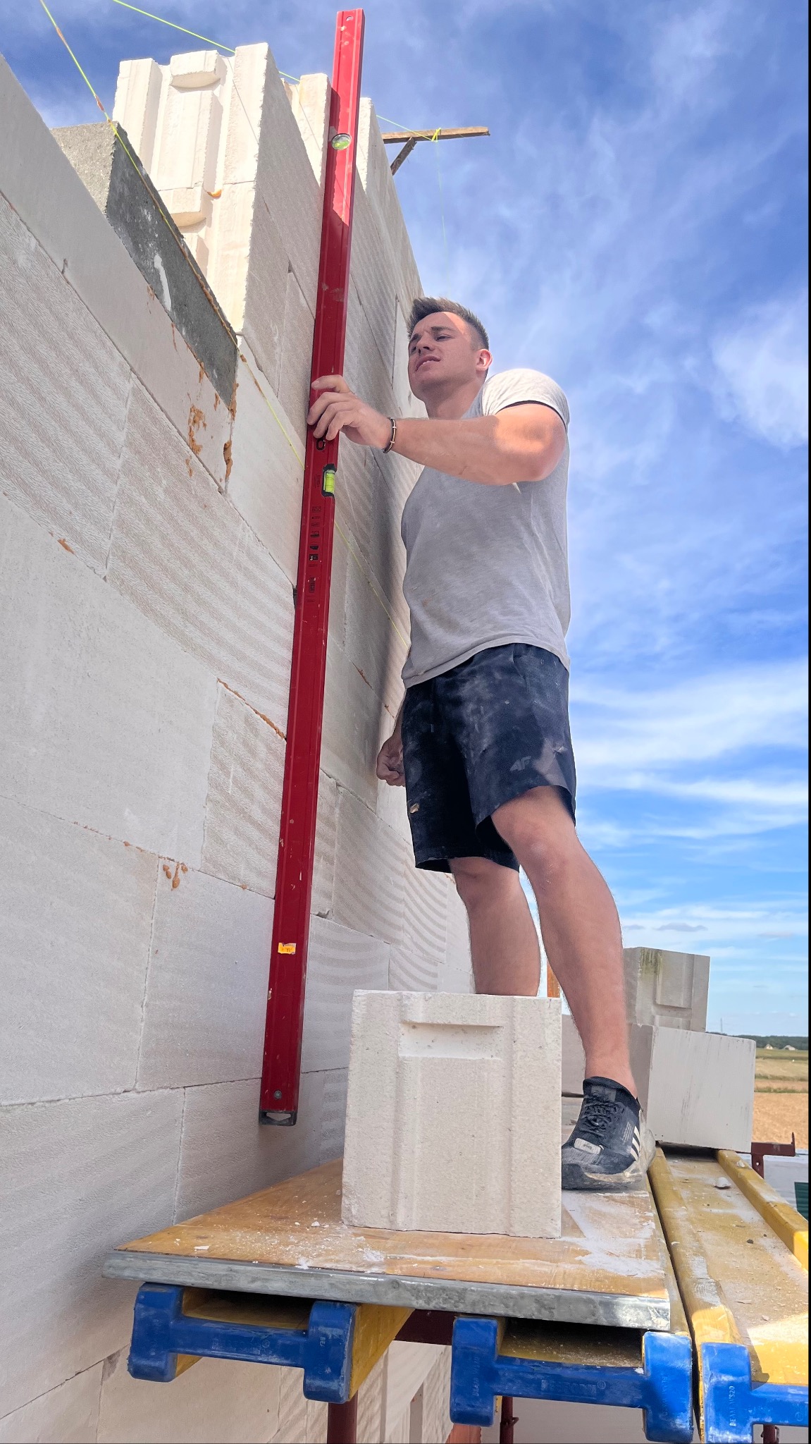 Mężczyzna na rusztowaniu, sprawdzający pionowość ściany z betonu komórkowego za pomocą poziomicy, widok z dołu na tle błękitnego nieba.