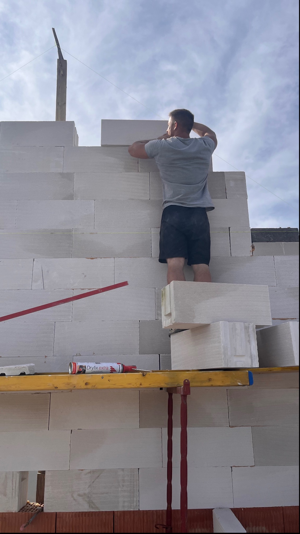 Murarz w szarej koszulce i czarnych spodenkach układa białe bloczki z betonu komórkowego na budowie, stojąc na rusztowaniu z poziomnicą i tubą kleju Dryfix extra, widok z dołu na niebo.