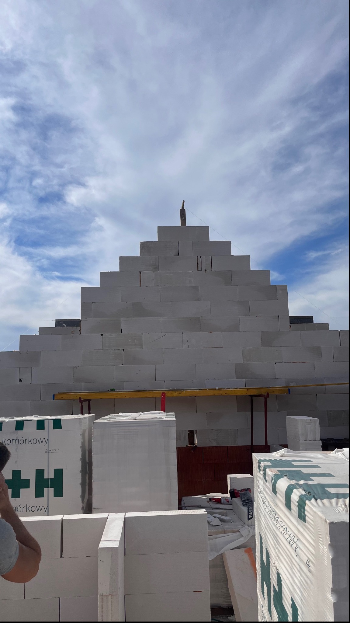 Wznoszona ściana z białych bloczków komórkowych o piramidalnym kształcie, z widocznym poziomowaniem i paletami materiałów budowlanych u podstawy, na tle błękitnego nieba.