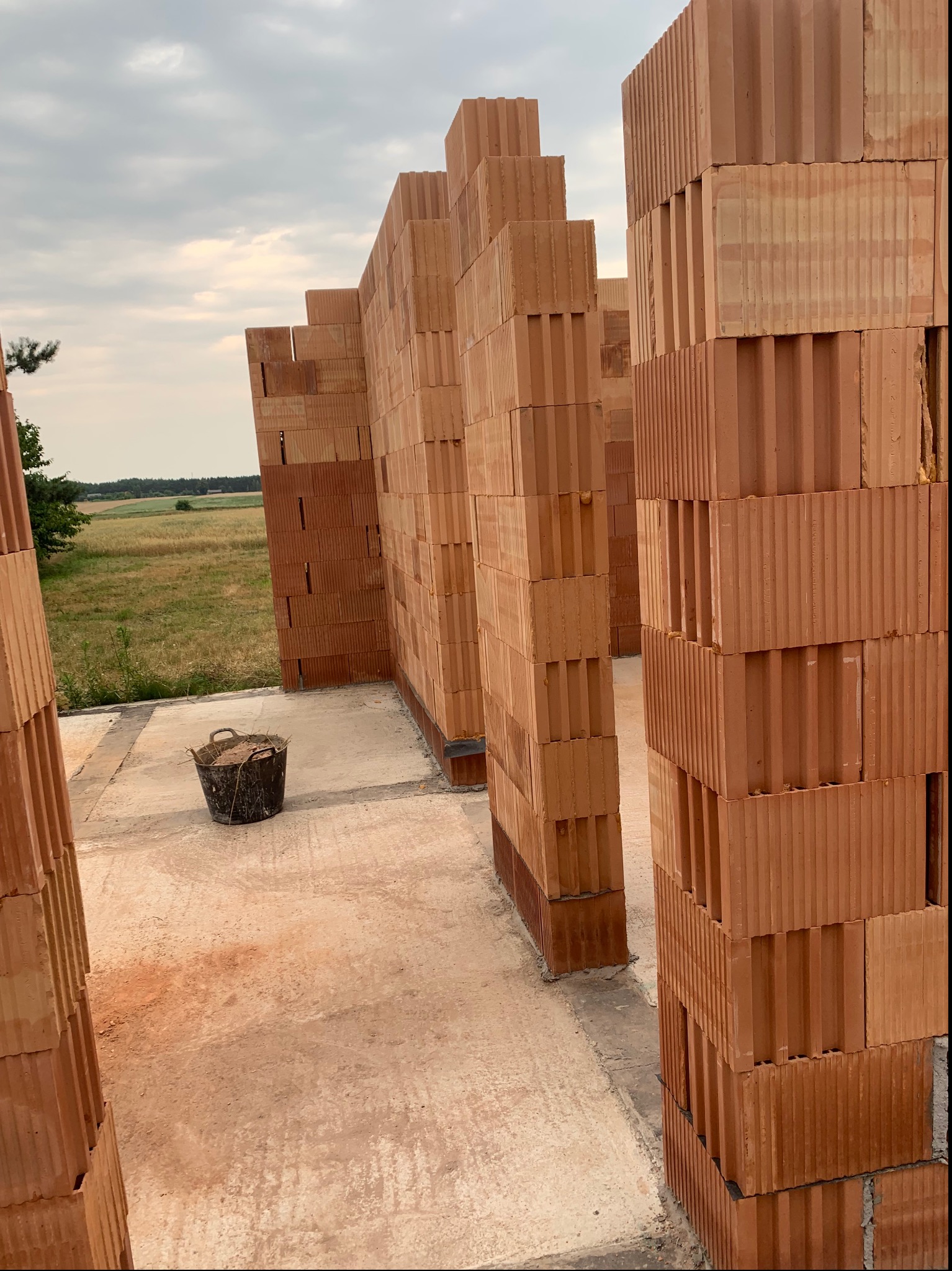 Wznoszone ściany z czerwonych bloczków ceramicznych na fundamencie, widoczne wiadro z resztkami zaprawy i otwarty krajobraz w tle.