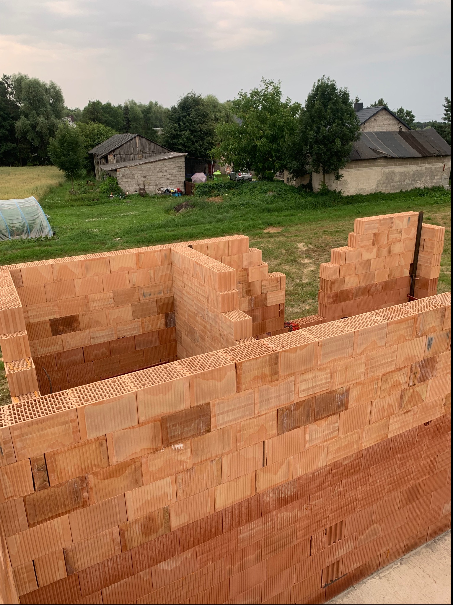 Wznoszenie ścian domu z czerwonych bloczków ceramicznych Porotherm na wsi, widoczne fragmenty muru z otworami, w tle zabudowania gospodarcze i zieleń.