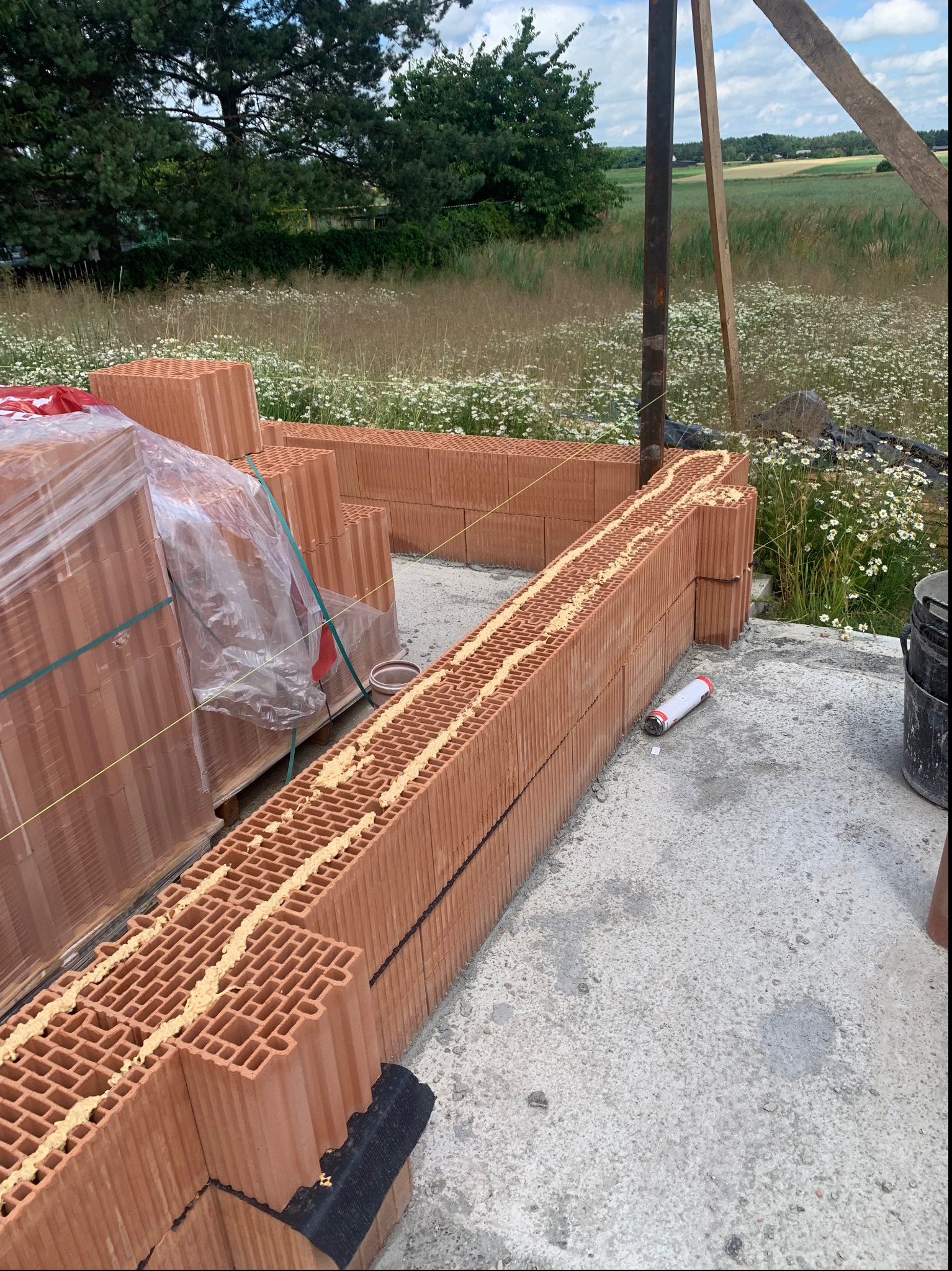 Wznoszenie muru z czerwonej cegły ceramicznej Porotherm na zaprawie klejowej, widoczne puszki kleju, w tle łąka z kwitnącymi kwiatami i drzewami.