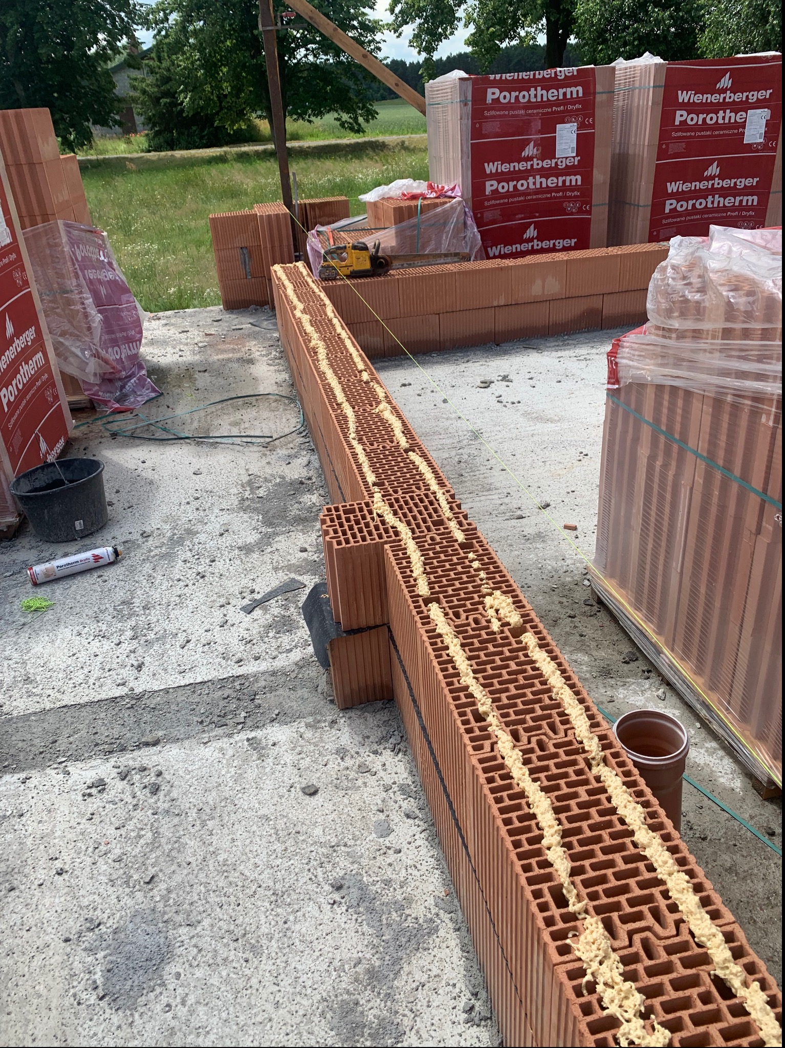 Wznoszenie ściany z pustaków ceramicznych Porotherm Dryfix, z widoczną warstwą zaprawy klejowej w spoinach poziomych, na tle palet z zapakowanymi pustakami.