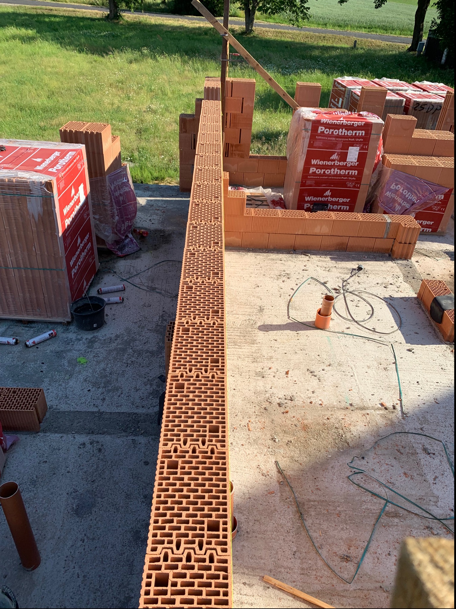 Wznoszenie muru z pustaków ceramicznych Porotherm na budowie, widoczne palety z zapakowanymi pustakami i narzędzia budowlane na betonowej posadzce.
