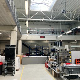 Architektura Będkowscy sp. z o.o. - Wnętrze hali produkcyjnej z antresolą, widoczne regały, wózki transportowe i pracownik na górnym poziomie.