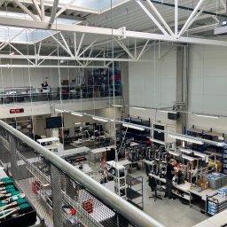Architektura Będkowscy sp. z o.o. - Wnętrze hali produkcyjnej z widokiem z góry na stanowiska pracy, gdzie pracownicy montują i testują urządzenia elektryczne; widoczna konstrukcja stalowa dachu i regały z komponentami.