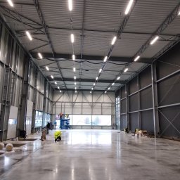 Architektura Będkowscy sp. z o.o. - Wnętrze hali produkcyjnej w trakcie budowy, widoczne stalowe elementy konstrukcyjne, oświetlenie LED pod sufitem, pracownicy na podnośniku oraz robotnicy przy posadzce.