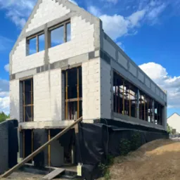 Surowy stan dwupiętrowego domu w trakcie budowy z białych bloczków, widoczne żółte podpory okien, czarna folia fundamentowa i niebo z chmurami w tle.