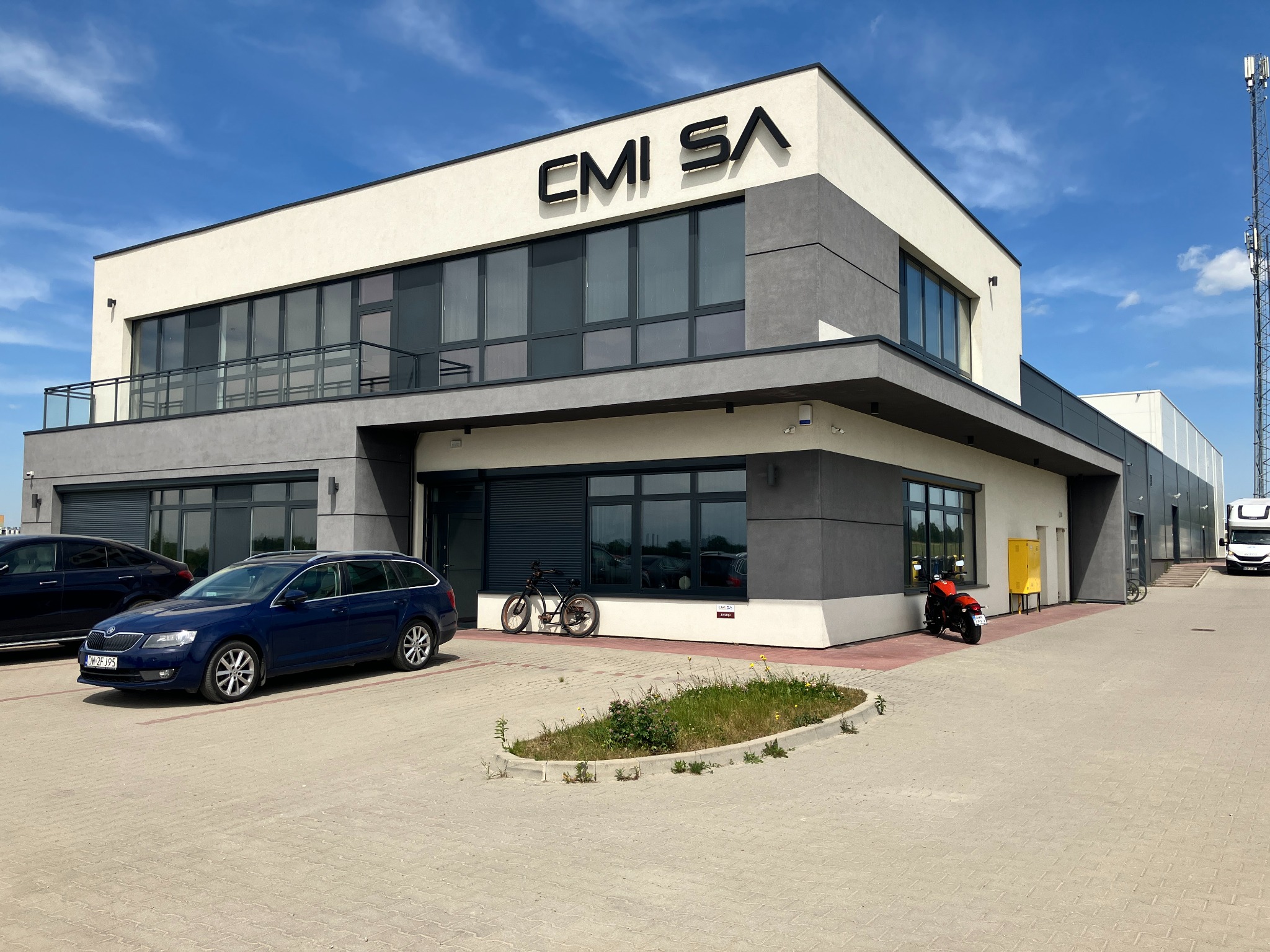 Nowoczesny budynek biurowy z logo CMI SA, szaro-biała elewacja, duże okna, zaparkowane samochody i motocykl przed budynkiem, błękitne niebo.