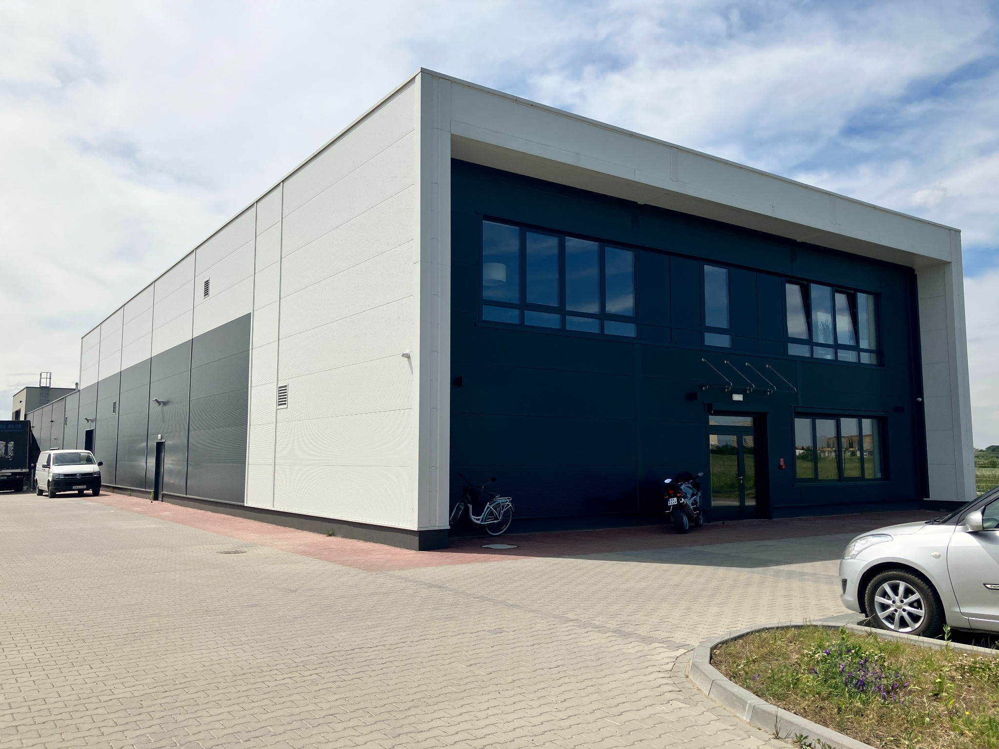 Nowoczesny budynek usługowy z dwoma motocyklami i rowerem zaparkowanymi przed wejściem, fasada w kolorach białym i grafitowym.