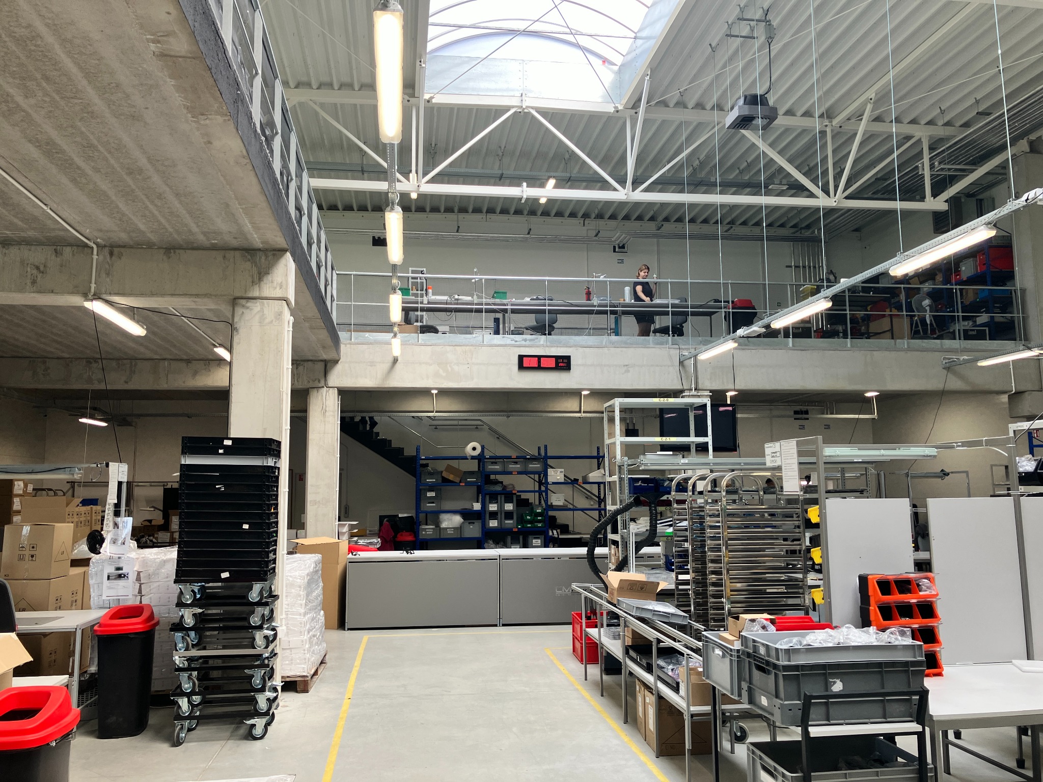 Wnętrze hali produkcyjnej z antresolą, widoczne regały, wózki transportowe i pracownik na górnym poziomie.