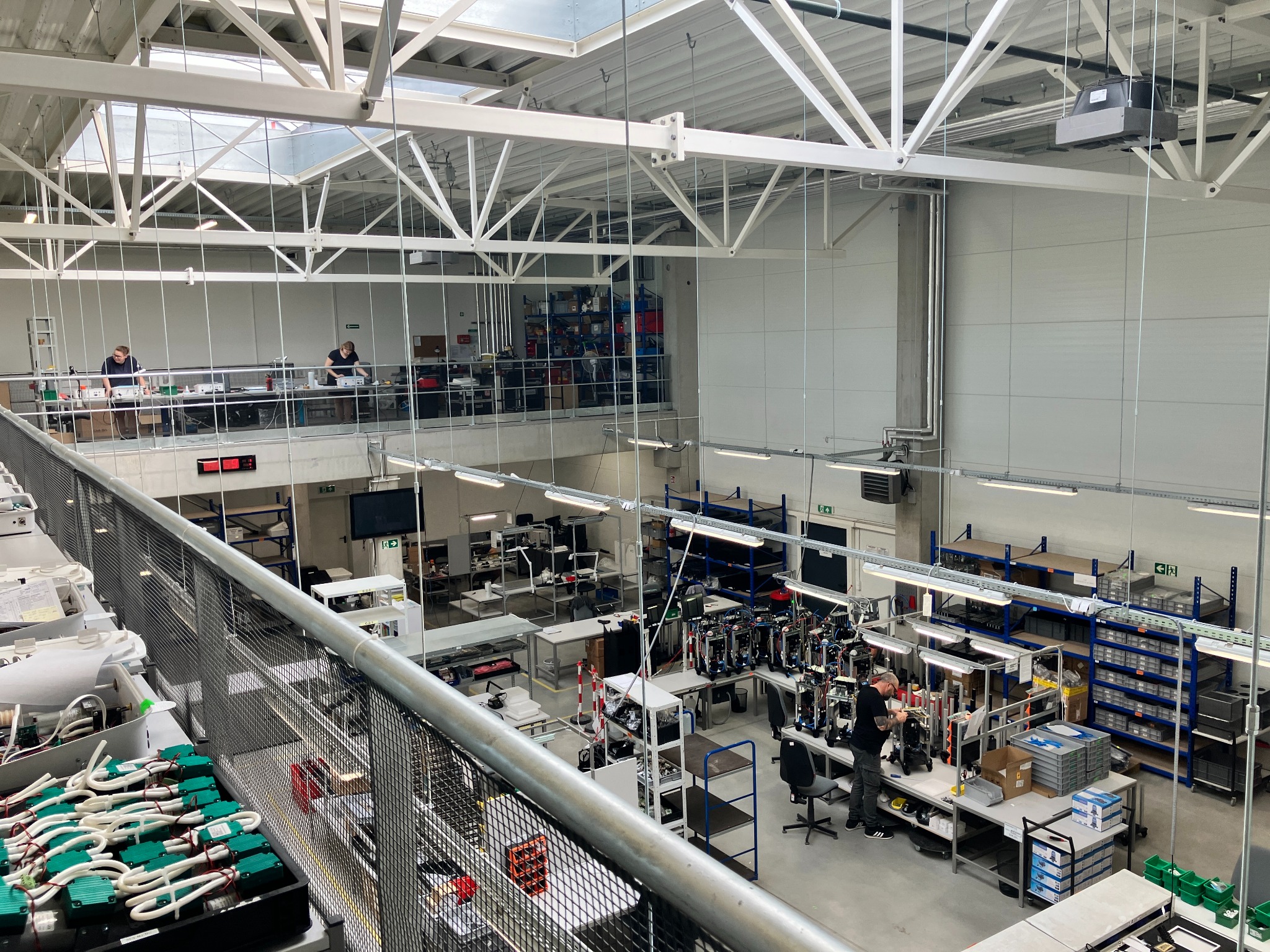 Wnętrze hali produkcyjnej z widokiem z góry na stanowiska pracy, gdzie pracownicy montują i testują urządzenia elektryczne; widoczna konstrukcja stalowa dachu i regały z komponentami.