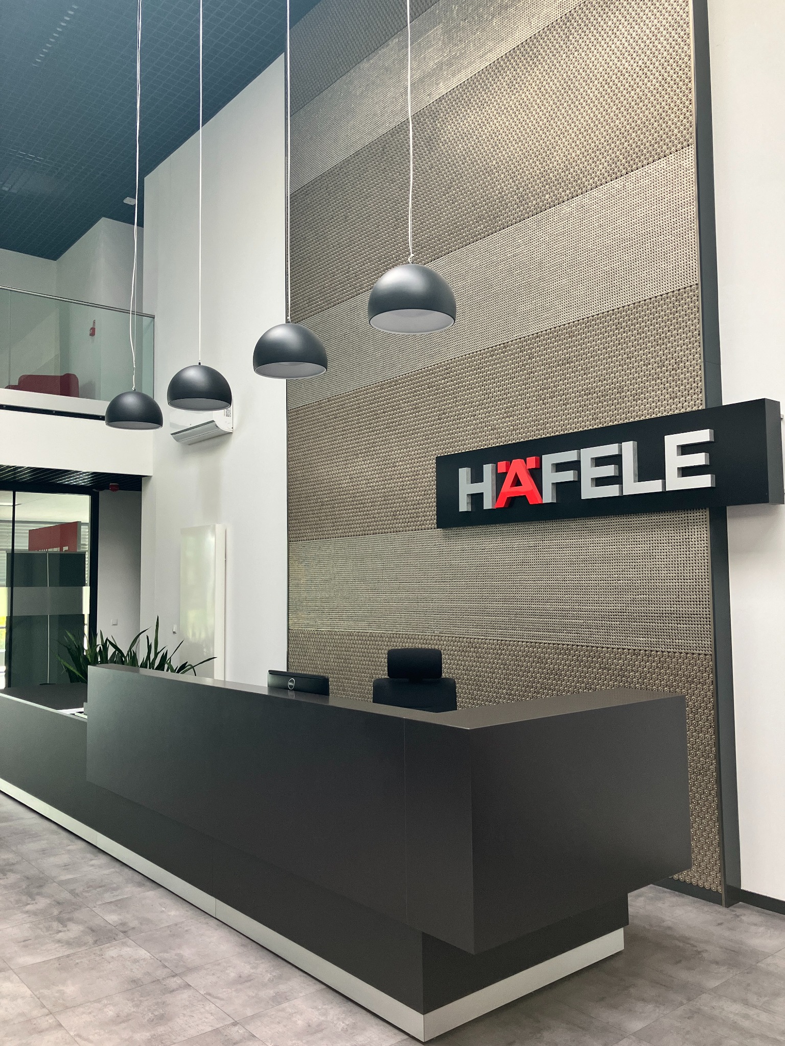 Recepcja w nowoczesnym biurze we Wrocławiu z ciemnoszarą ladą, trzema wiszącymi lampami i ścianą z dekoracyjnym panelem oraz logo firmy Häfele.