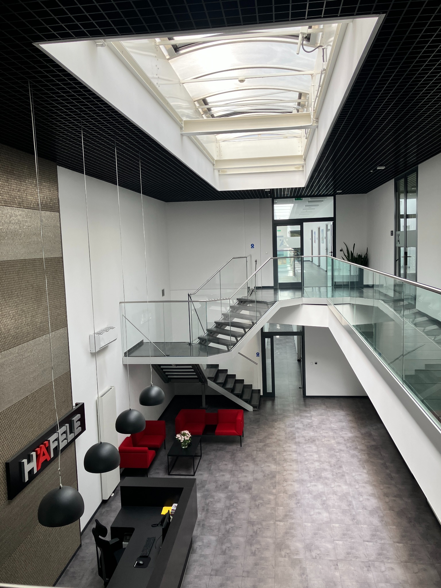 Wnętrze nowoczesnego biurowca z antresolą, przeszklonymi balustradami, czarnymi schodami, czerwonymi sofami w strefie relaksu i przeszklonym dachem z widokiem na niebo.