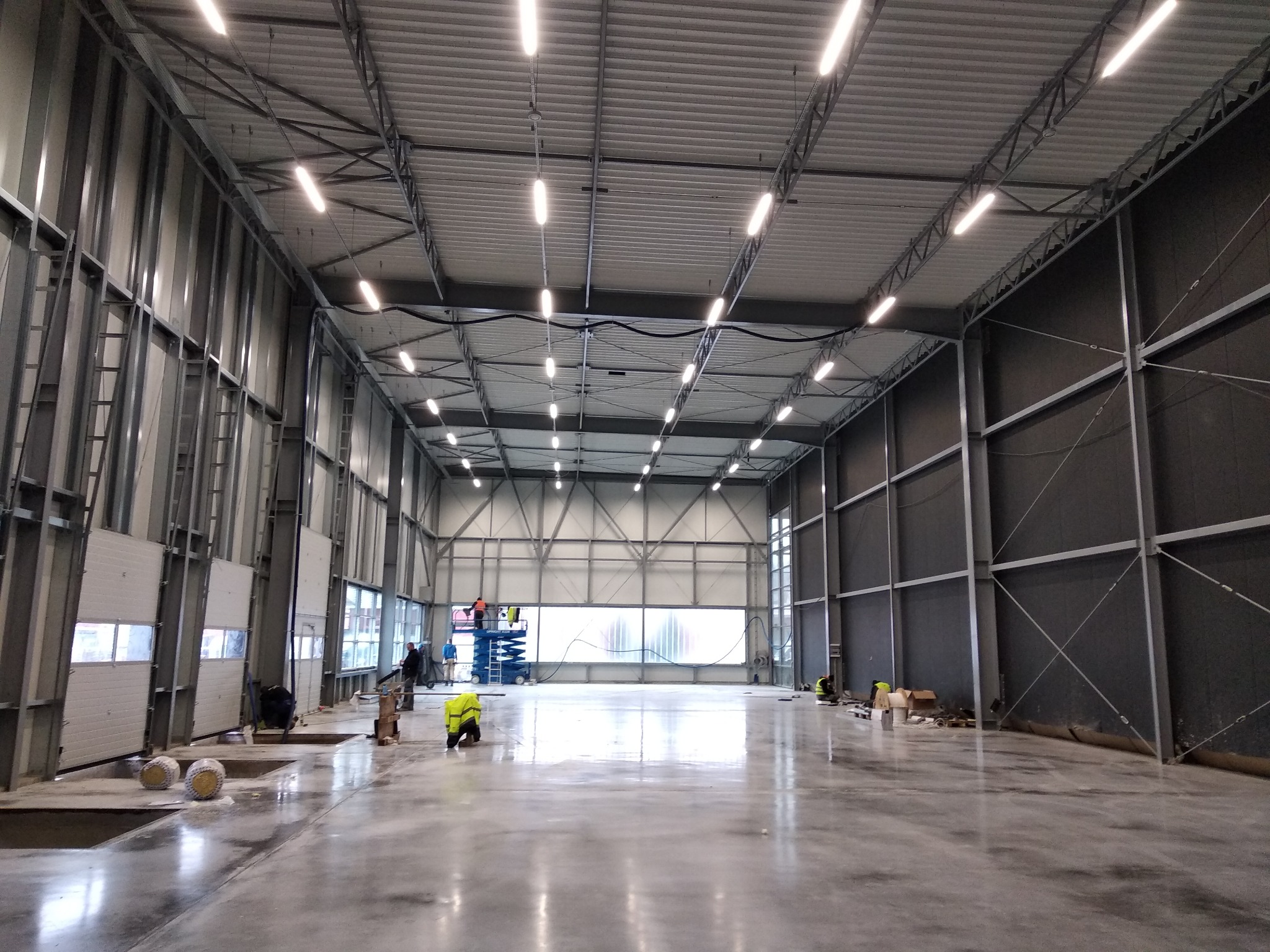 Wnętrze hali produkcyjnej w trakcie budowy, widoczne stalowe elementy konstrukcyjne, oświetlenie LED pod sufitem, pracownicy na podnośniku oraz robotnicy przy posadzce.