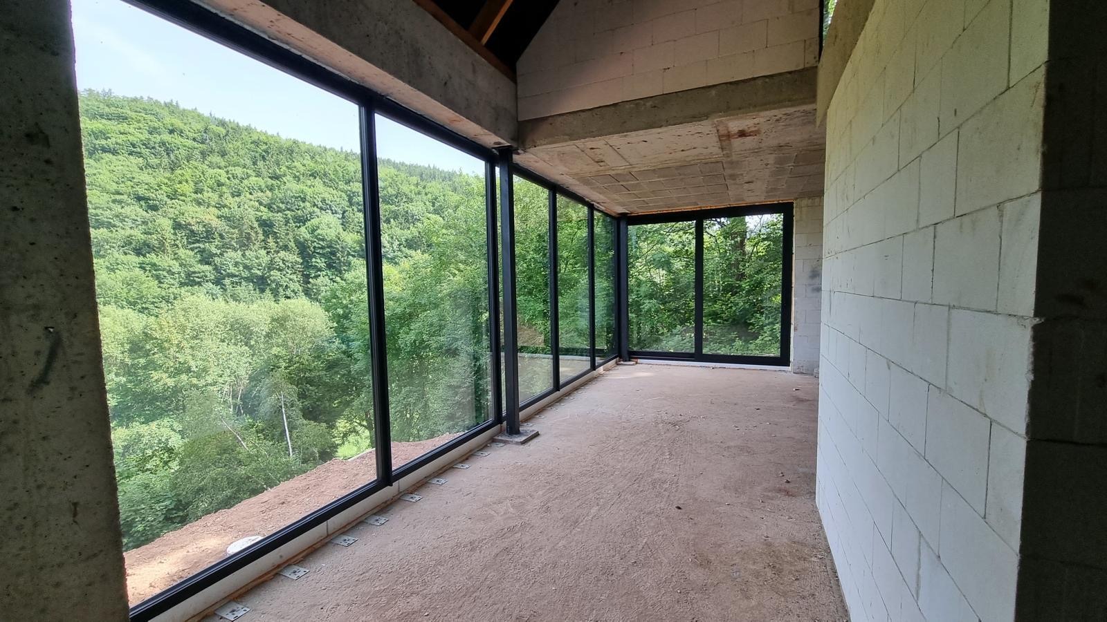 Wnętrze surowego domu z dużymi oknami z widokiem na las, ściany z betonu komórkowego, podłoga wylewka, widoczne elementy konstrukcyjne stropu.