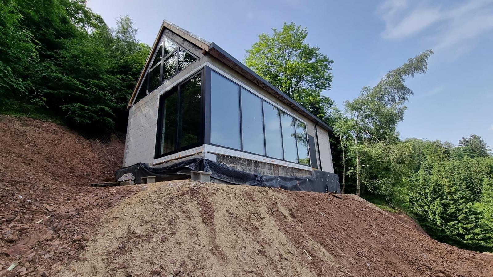 Budynek w trakcie budowy z dużymi oknami, usytuowany na wzgórzu, otoczony zielenią.