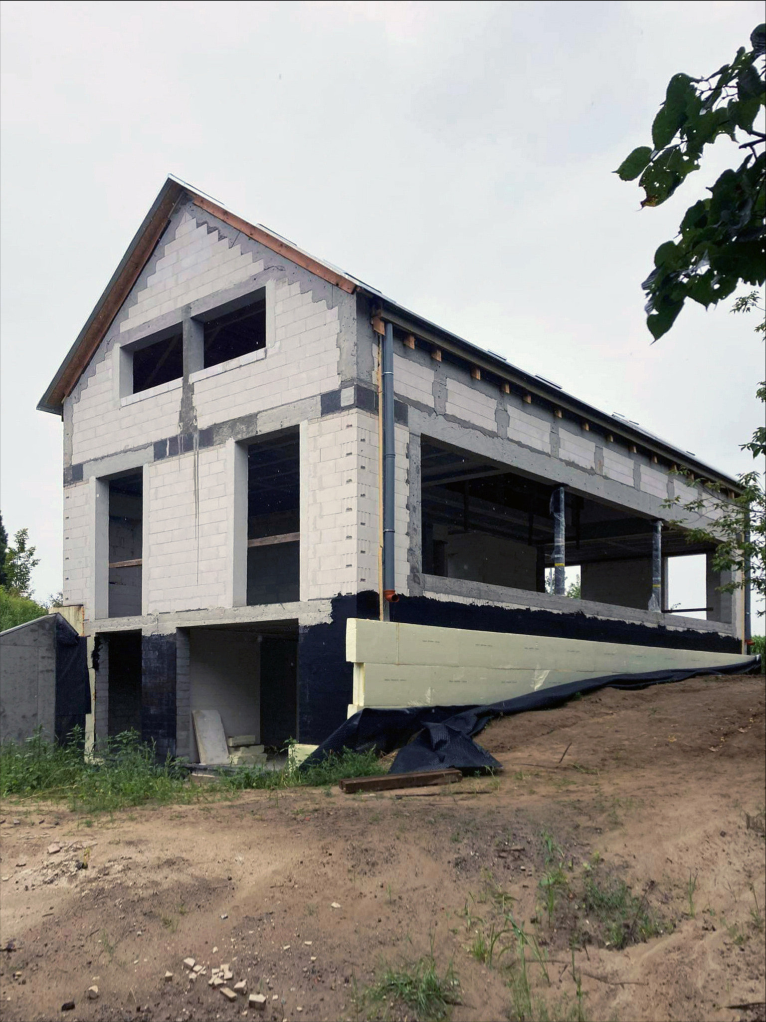 Dom w trakcie budowy z białych bloczków, widoczne okna bez szyb, ocieplenie fundamentów i teren budowy.