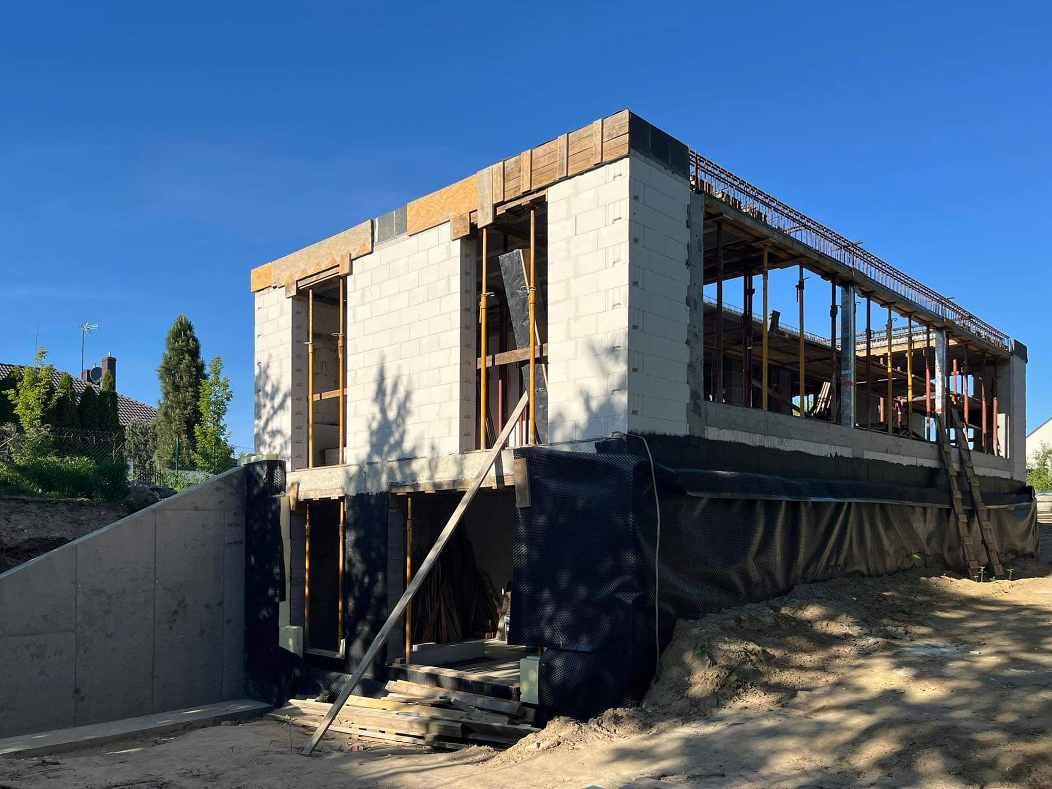 Surowy stan budynku jednorodzinnego w trakcie budowy, widoczne ściany z bloczków, szalunki stropu i zabezpieczenie fundamentów folią kubełkową.