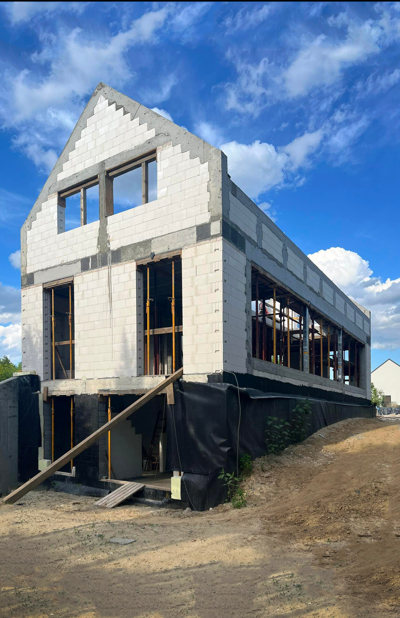 Surowy stan dwupiętrowego domu w trakcie budowy z białych bloczków, widoczne żółte podpory okien, czarna folia fundamentowa i niebo z chmurami w tle.