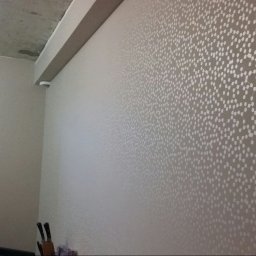 budobel.pl - Ściana w pokoju z nową tapetą w drobne białe kropki na szarym tle, widoczny fragment surowego betonowego sufitu i niezabezpieczony włącznik światła.