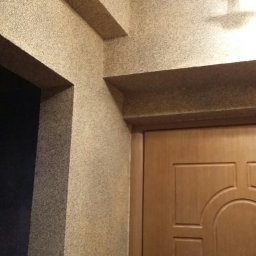 budobel.pl - Beżowa ściana z dekoracyjną strukturą, widoczne elementy instalacji elektrycznej pod sufitem i fragment drewnianych drzwi z ozdobnym frezowaniem.