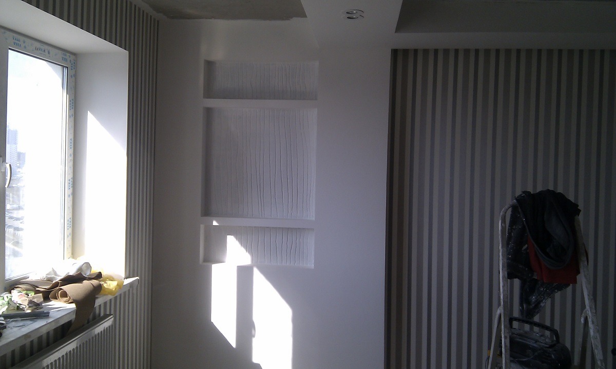 Wnętrze pokoju w trakcie prac wykończeniowych: widok na okno z materiałami na parapecie, wnękę w ścianie z fakturą oraz fragment ściany w pionowe pasy z drabiną opartą o nią.