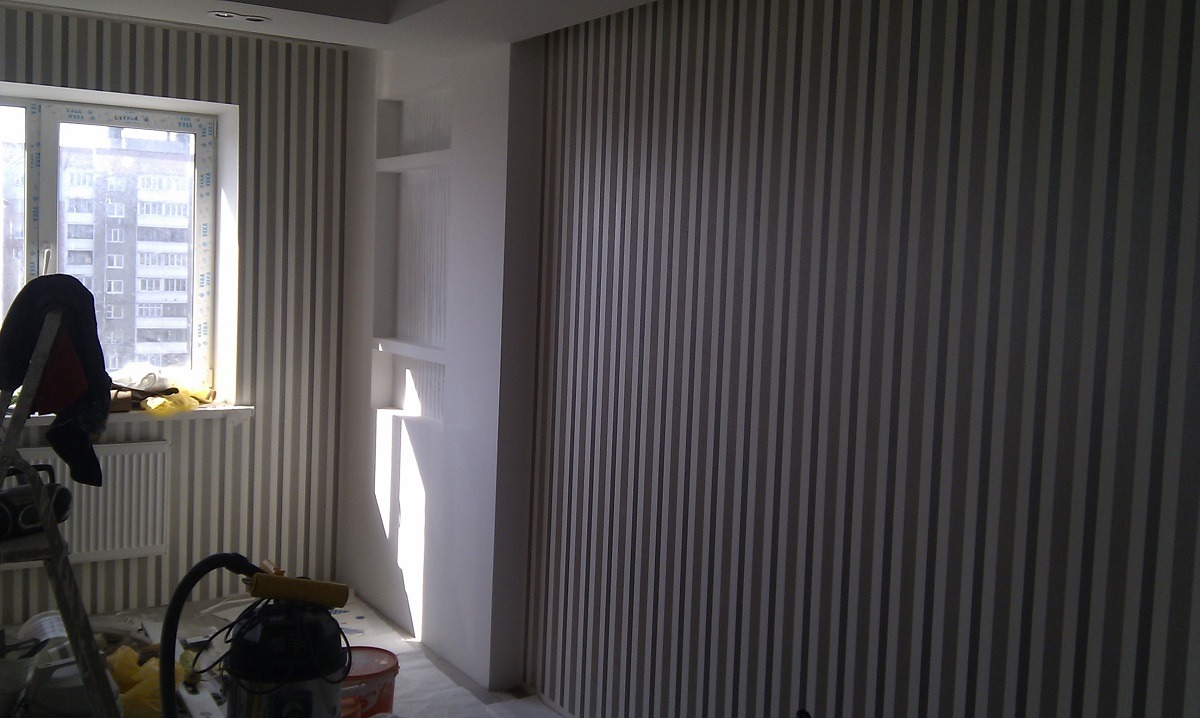 Pionowe pasy w odcieniach szarości na ścianie w pokoju, widoczny fragment okna z widokiem na blok, drabina malarska z narzędziami i akcesoriami malarskimi.
