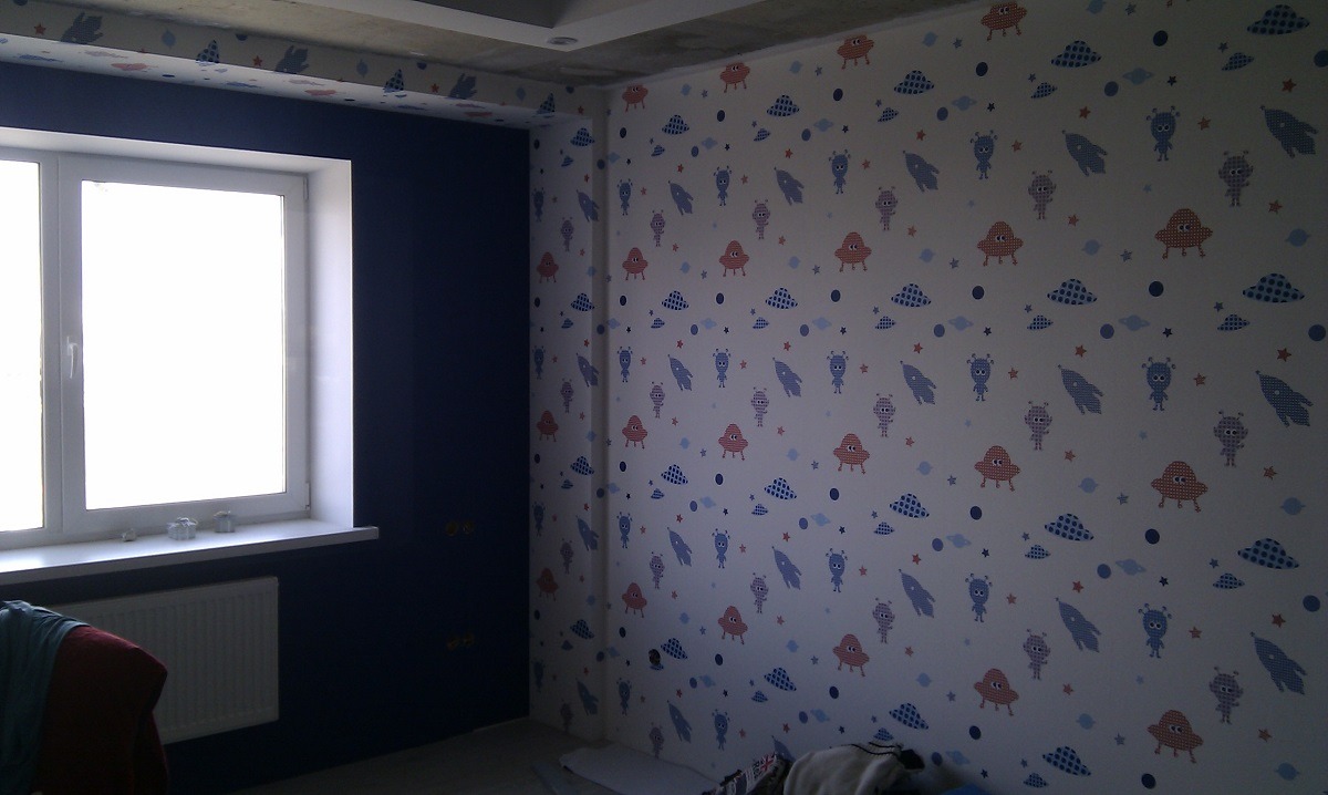 Pokój dziecięcy po remoncie: ściany pokryte tapetą w kosmiczne motywy (statki kosmiczne, ufoludki), jedna ściana pomalowana na granatowo, widoczne okno z białymi ramami i grzejnik.
