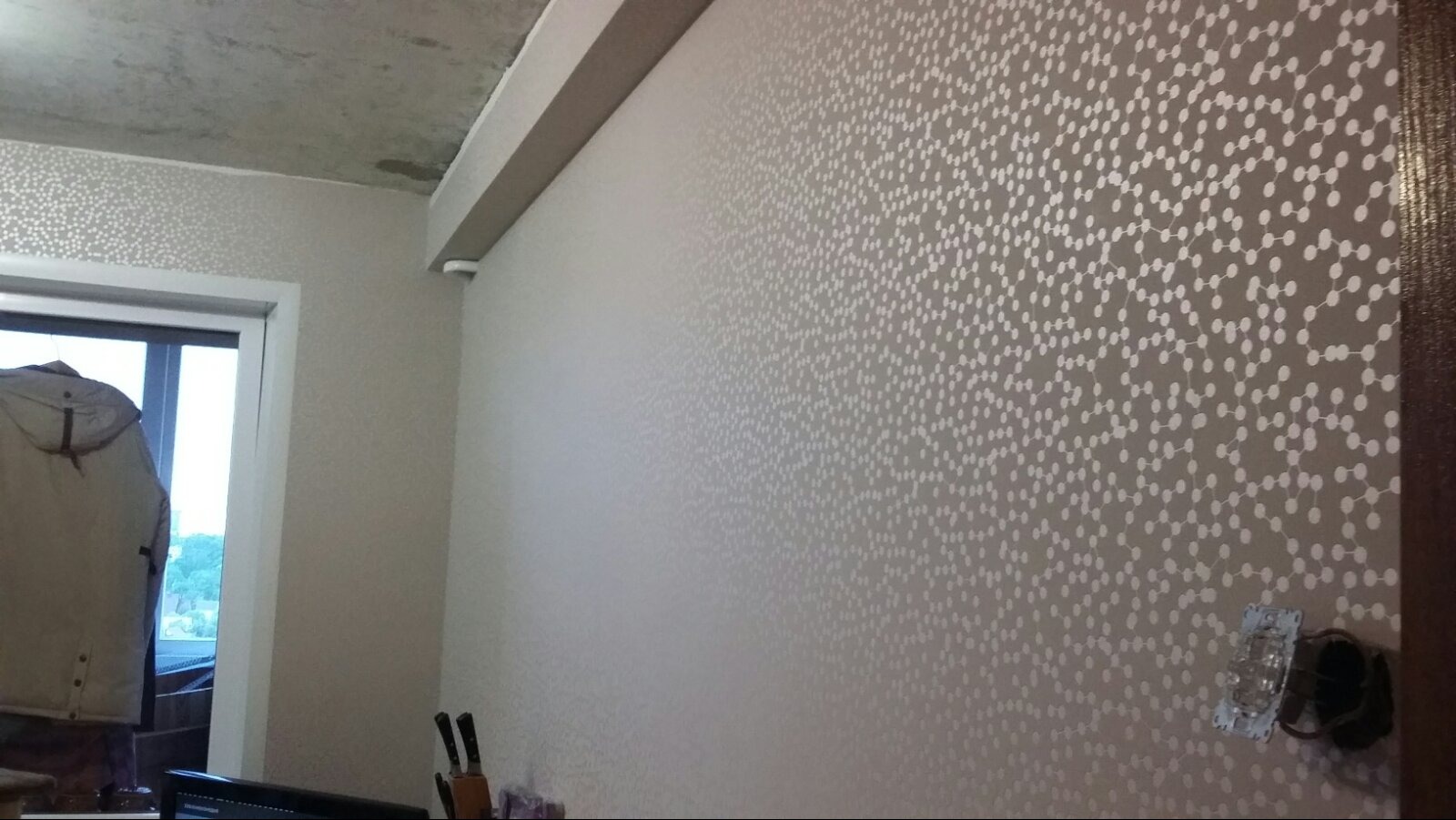 Ściana w pokoju z nową tapetą w drobne białe kropki na szarym tle, widoczny fragment surowego betonowego sufitu i niezabezpieczony włącznik światła.