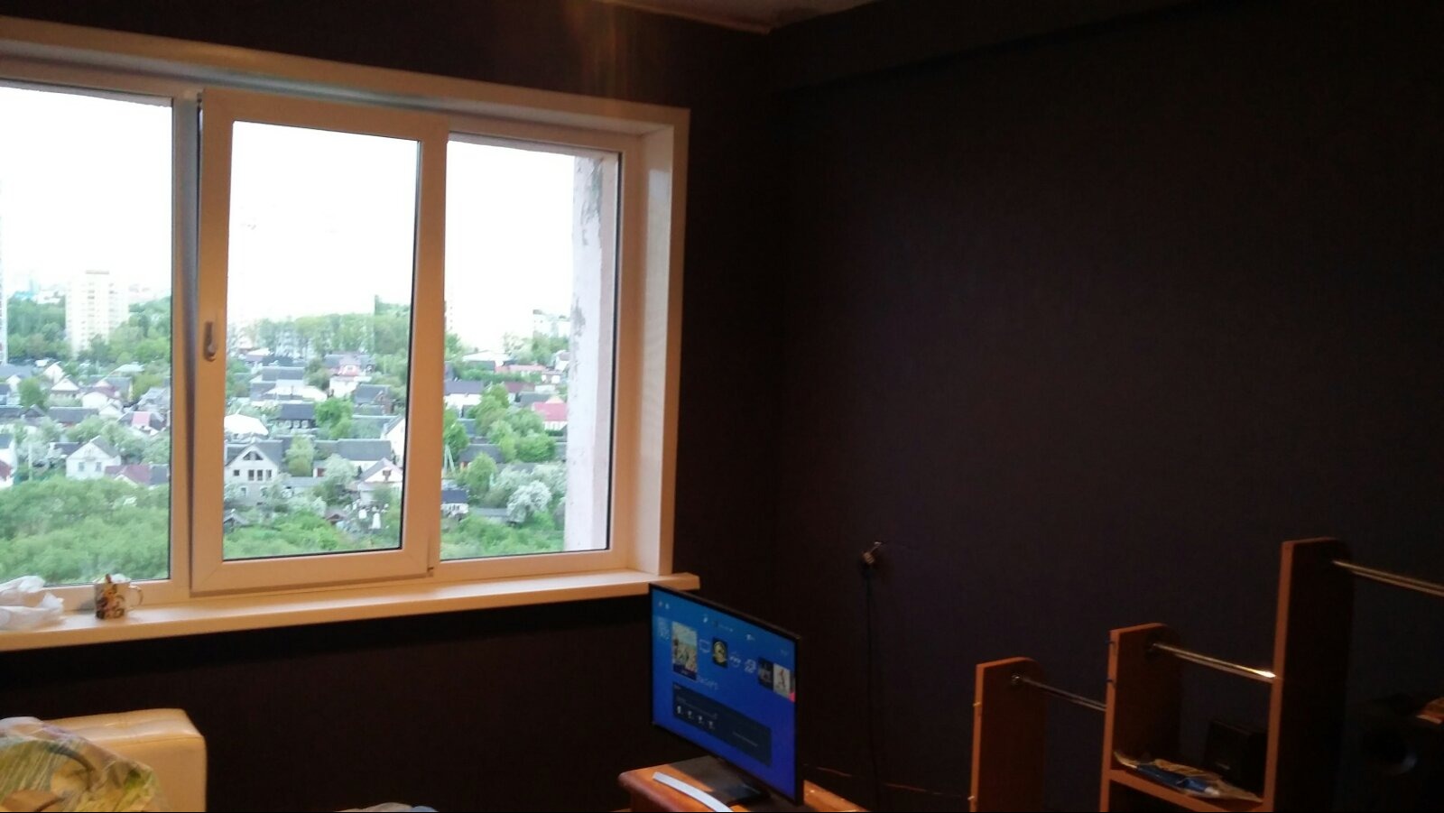 Pokój po malowaniu na ciemny kolor z widokiem na osiedle domów jednorodzinnych za oknem; widoczny telewizor i regał.