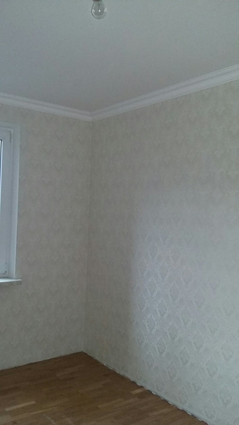 Pomalowane wnętrze pokoju z wzorzystą tapetą w odcieniach bieli, drewnianą podłogą i listwami przypodłogowymi, oświetlone pojedynczą żarówką.