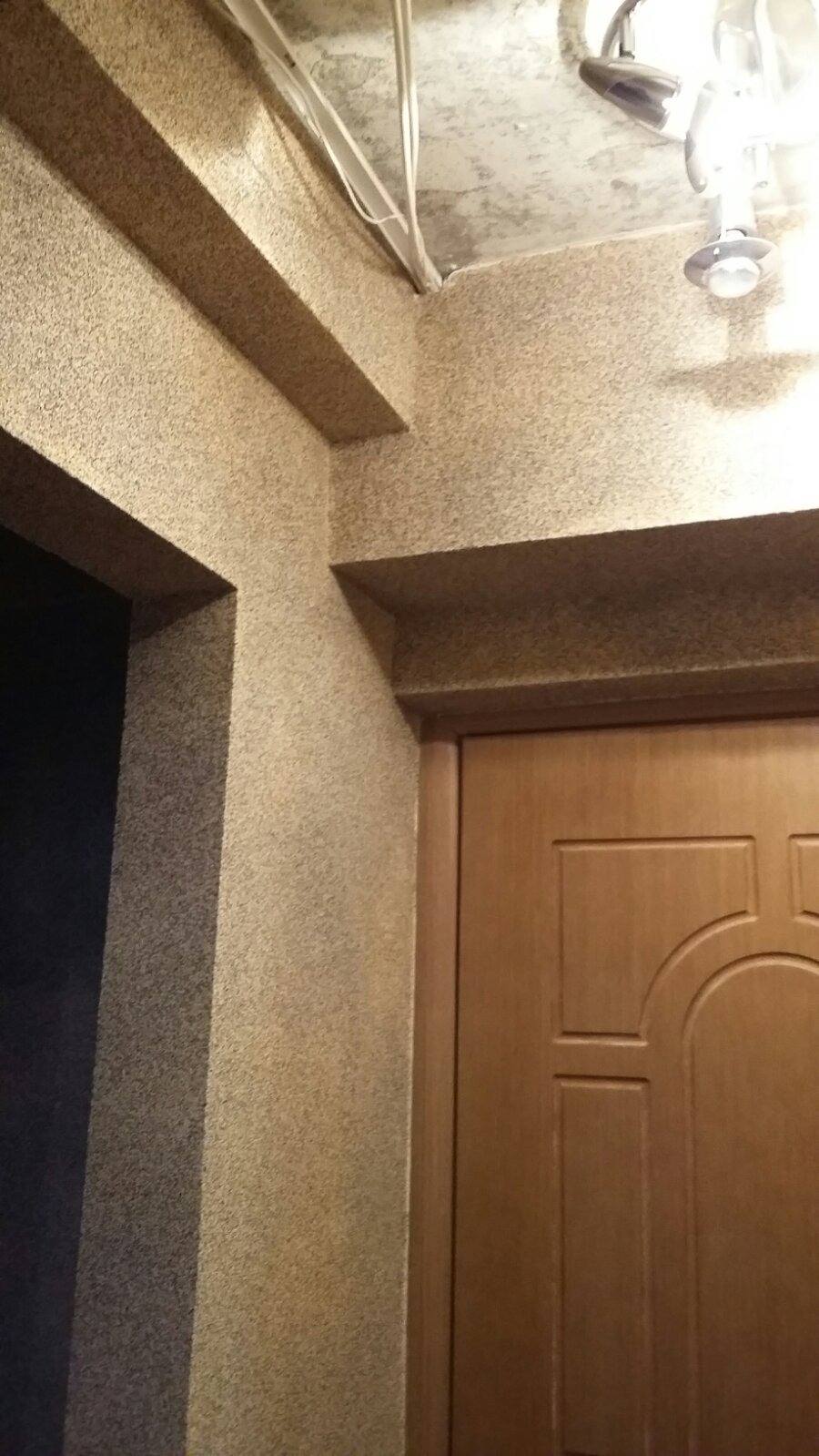 Beżowa ściana z dekoracyjną strukturą, widoczne elementy instalacji elektrycznej pod sufitem i fragment drewnianych drzwi z ozdobnym frezowaniem.