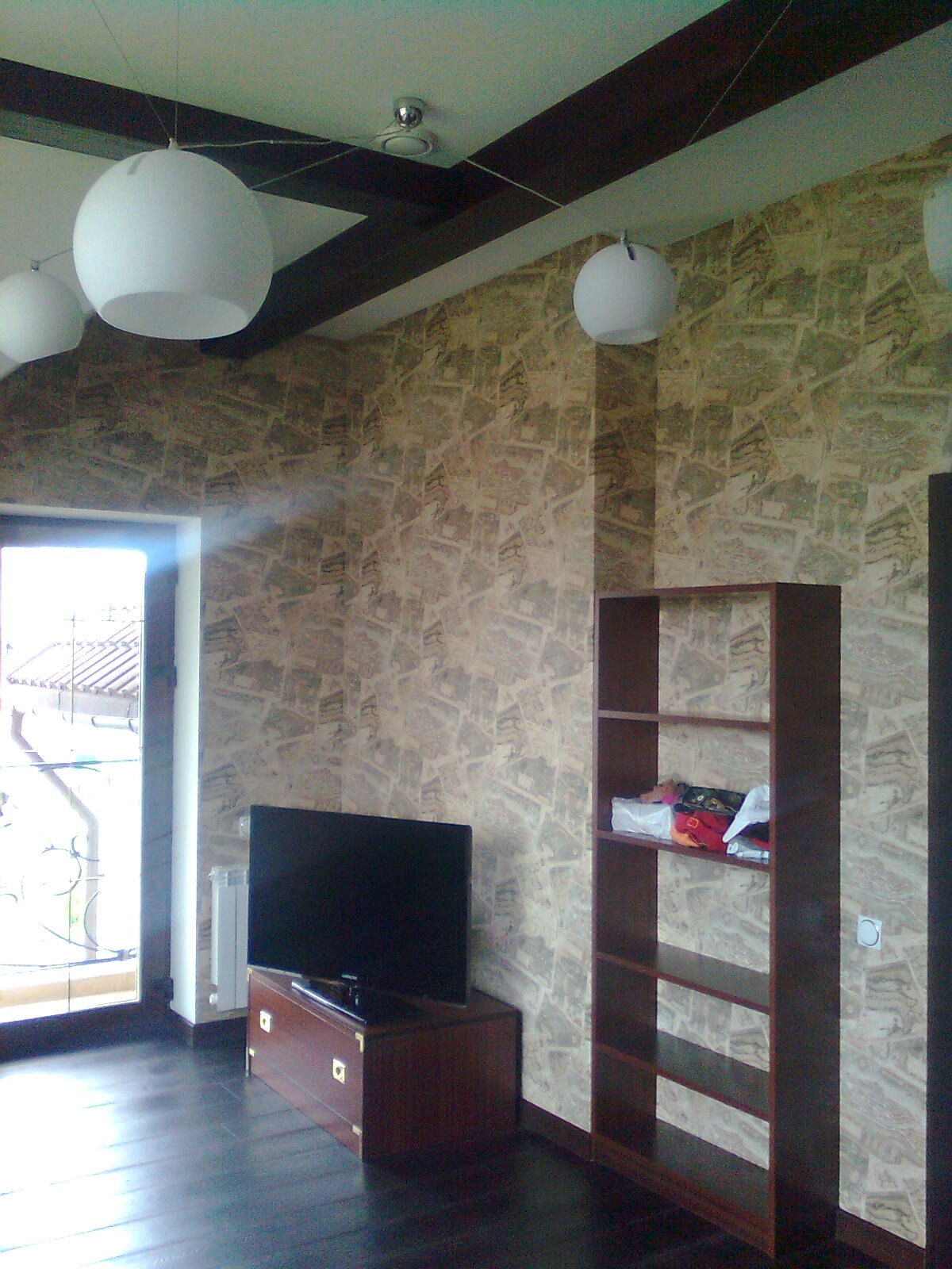 Pokój po remoncie: ściana pokryta wzorzystą tapetą w odcieniach beżu i brązu, ciemna podłoga, drewniane belki na suficie, białe, okrągłe lampy wiszące, ciemne meble, widoczne drzwi balkonowe.