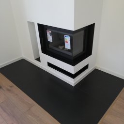 AKABUD - Nowoczesny narożny kominek z czarnym wkładem i białym obramowaniem, widoczne naklejki informacyjne na szybie, czarna podłoga z płytek kontrastująca z drewnianą podłogą w jodełkę.