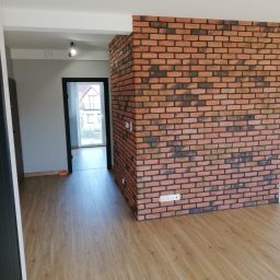AKABUD - Wnętrze mieszkania z wykończoną ścianą z cegły klinkierowej, widoczna drewnopodobna podłoga i otwarte przejście do kolejnego pomieszczenia z ościeżnicą w kolorze antracytowym.