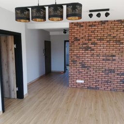 AKABUD - Nowoczesne wnętrze po remoncie: parkiet laminowany w odcieniach beżu, ściana z czerwonej cegły, czarne lampy sufitowe z ażurowymi kloszami i czarne drzwi z drewnianym fornirem.